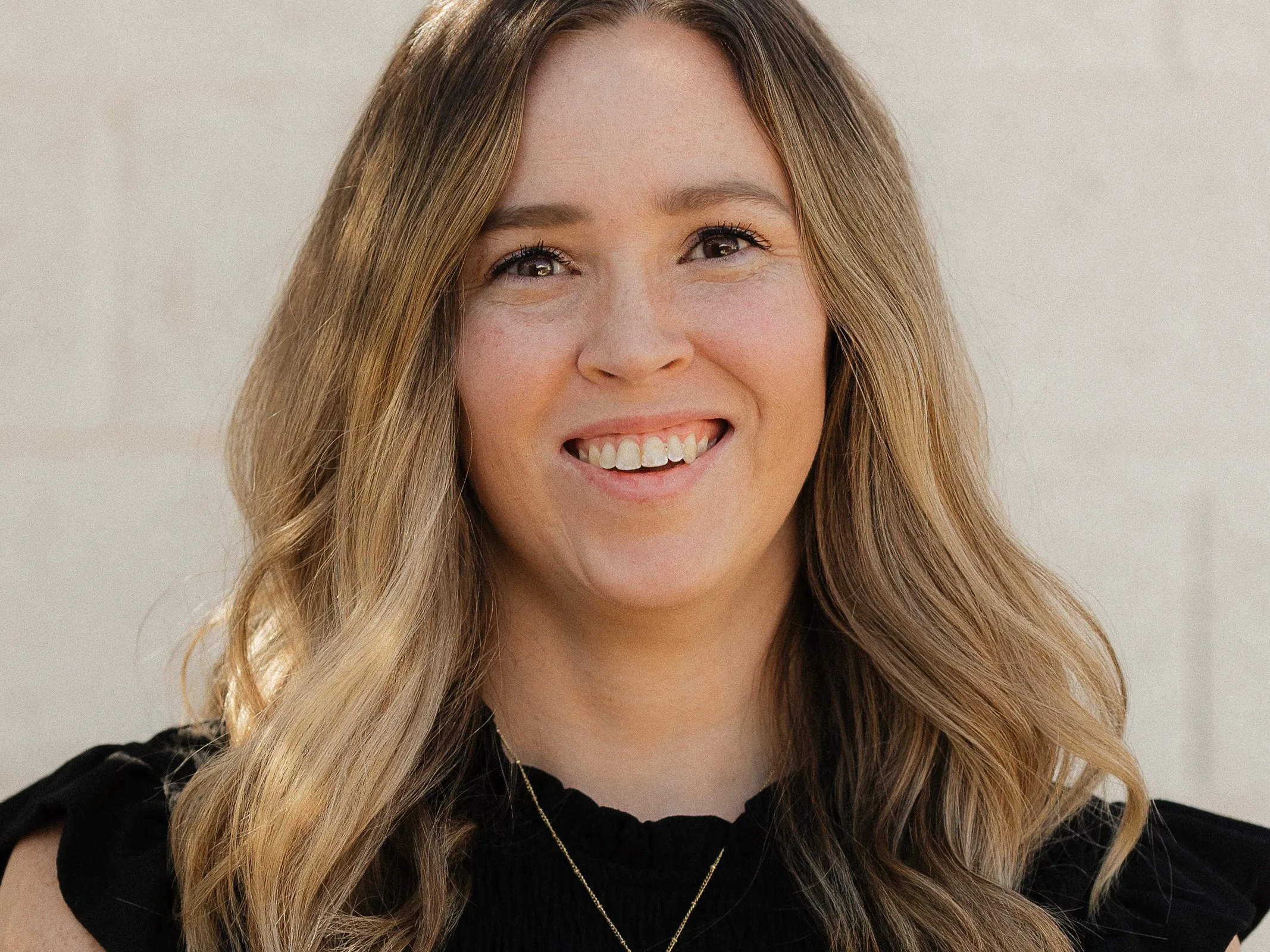 Megan Riddle smiling with her arms crossed, in a black top against a light block wall